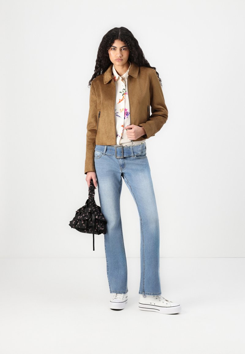Giacca in suede marrone sopra una camicia a fiori con bottoni, jeans a campana blu e sneakers bianche, mentre tiene una piccola borsa nera con dettagli floreali.
