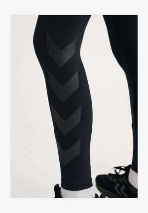 Zwarte leggings met een glanzend chevronpatroon langs het onderbeen. Gemaakt van glad, rekbaar materiaal. Gecombineerd met zwarte sportschoenen.