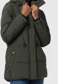 Veste matelassée vert foncé avec une capuche, fermée par une fermeture éclair et des boutons. Comprend des cordons élastiques à la taille et des accents aux poignets.