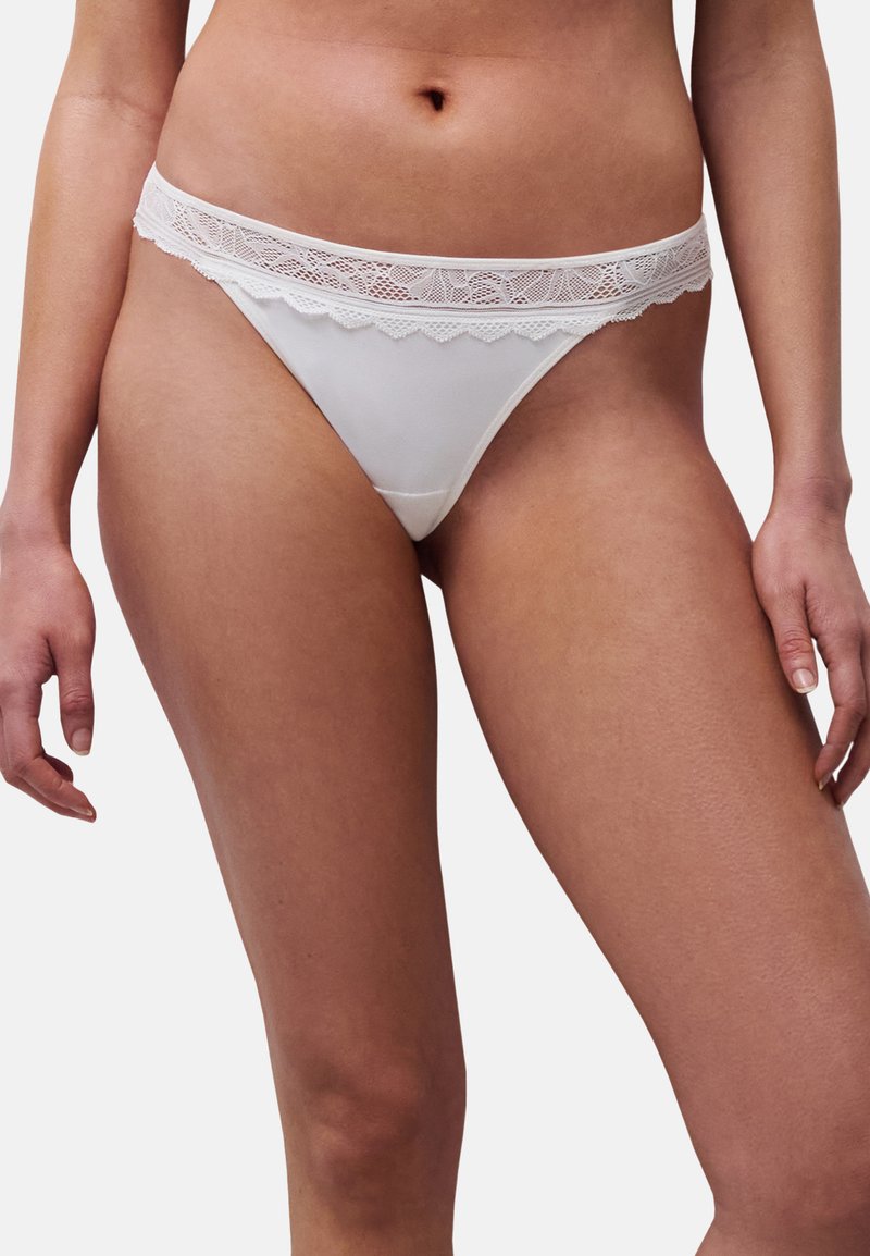Frau trägt Unterwäsche im Bikini-Stil mit Spitze, die den Oberkörper und die Oberschenkel zeigt, vor einem einfarbigen Hintergrund.