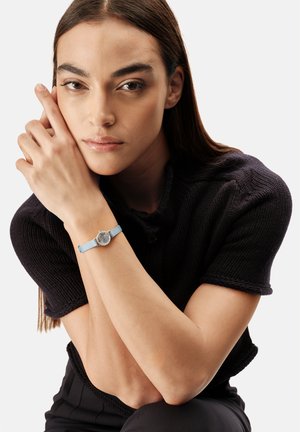 Kvinde med langt brunt hår iført en sort strikket top med korte ærmer, der hviler sit ansigt på sine korsede hænder med en lyseblå armbåndsur.