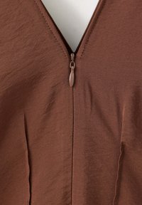 Gros plan d’un vêtement marron avec une fermeture éclair frontale partiellement fermée, montrant la texture du tissu et les détails de couture.