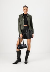 Olivengrønn semsket jakke, svart crop top, svarte skinnlignende shorts, svart håndveske, kne-høye svarte støvler med spisse tær og spenner.