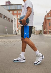 Idrottsskor med en vit, grå och svart design, texturerad ovandel, vadderad krage och ljusblå sula. Används med svarta shorts och en vit skjorta.