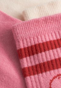 Gros plan sur des chaussettes tricotées roses avec deux rayures horizontales rouges sur une paire et une chaussette tricotée crème unie en arrière-plan.