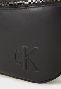 Borsa in pelle nera con logo "cK" in rilievo e zip visibile lungo il bordo superiore.