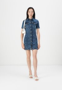 Robe chemise en denim, manches courtes, devant à boutons, deux poches poitrine, délavage clair, ourlet légèrement effiloché, associée à des sandales à talons blocs blanches.
