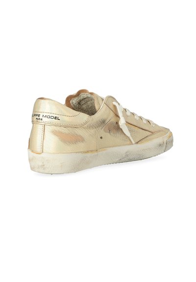 Sneaker basso in oro usurato con lacci bianchi e segni di usura visibili, angolato per mostrare il lato esterno e il retro con il logo del marchio sul tallone.
