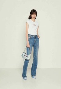 White fitted T-shirt with a cutout detail, paired with light blue flared jeans. Holding a small iridescent bag and wearing matching blue heels.