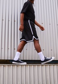 Shorts de sport noirs avec des rayures blanches, t-shirt noir, chaussettes blanches et baskets Converse noires, marchant devant un fond en métal ondulé.