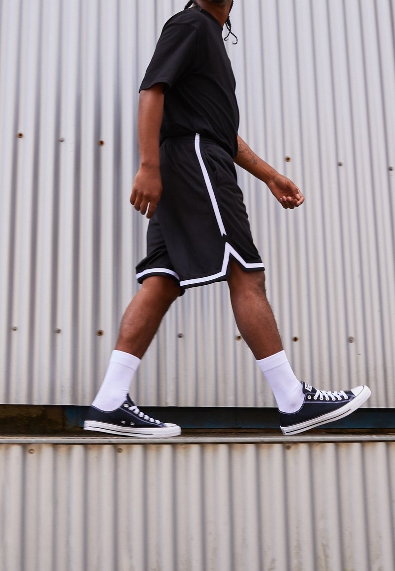 Shorts de sport noirs avec des rayures blanches, t-shirt noir, chaussettes blanches et baskets Converse noires, marchant devant un fond en métal ondulé.