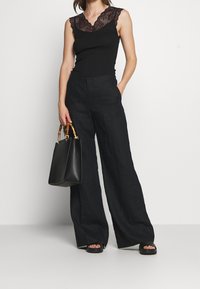 Svart ärmlös topp med spetskant som matchas med högmidjade svarta linnebyxor. En väldesignad svart handväska med bambuhandtag hålls.