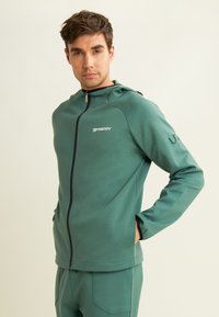 Hoodie zippé vert fabriqué en tissu doux et texturé. Il dispose de poches latérales et d'une capuche. Logo imprimé sur la poitrine et le bras.