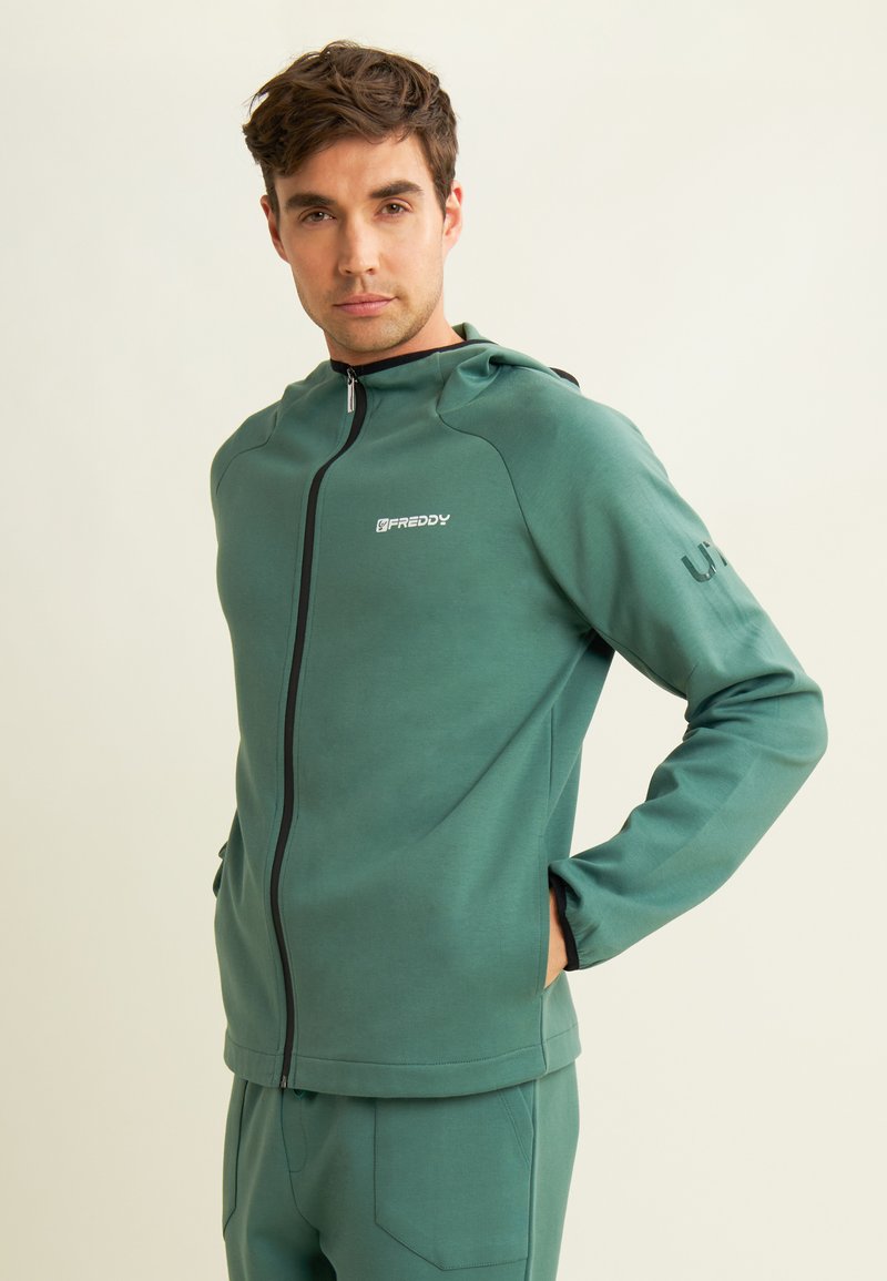 Hoodie zippé vert fabriqué en tissu doux et texturé. Il dispose de poches latérales et d'une capuche. Logo imprimé sur la poitrine et le bras.
