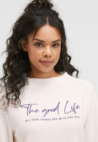 Helles rosa Sweatshirt mit Textur, das lila grafischen Text mit der Aufschrift "The good Life" und "ALLE GUTEN DINGE SIND WILDER UND FREI." zeigt.
