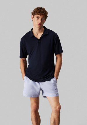 Polo shirt blu navy con colletto e chiusura a bottoni, abbinato a pantaloni corti grigio chiaro. Design semplice, texture simile al cotone e vestibilità rilassata.