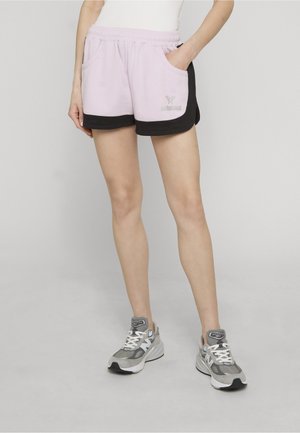 Frau trägt hellviolette Shorts mit schwarzem Besatz und graue Sportschuhe, steht mit einer Hand in der Tasche vor einem schlichten Hintergrund.