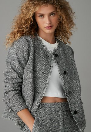 Getextureerde zwart-wit tweedjas met rafelranden, een ronde halslijn en zwarte knopen, gecombineerd met een witte cropped top.