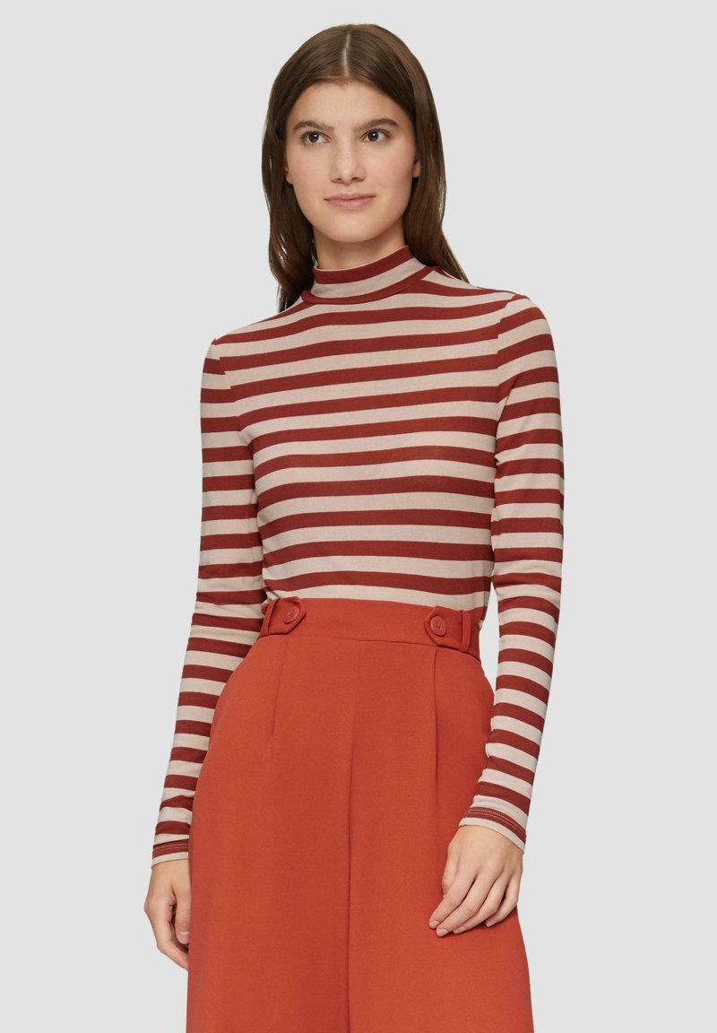 Pasiasty sweter z długim rękawem w kolorze burgundowym i beżowym, z wysokim kołnierzem. Połączony z pomarańczowymi spodniami w wysokim stanie z guzikami.