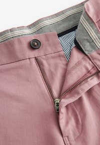 Gros plan sur un pantalon rose avec une fermeture éclair ouverte, un bouton noir, une bande de taille intérieure à motif visible et une doublure en tissu rayé.