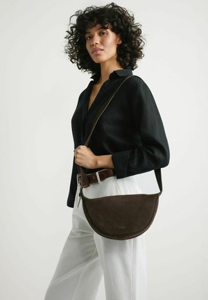 Kobieta o kręconych włosach ubrana w czarną koszulę i białe spodnie, trzymająca ciemnobrązową torbę na ramię z paskiem na ramię.