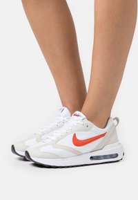 Białe i szare buty sportowe z pomarańczowym logo Nike, z teksturowaną górą z siateczki, amortyzowaną podeszwą i widoczną poduszką powietrzną.