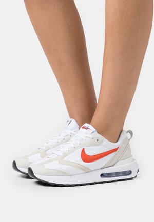 Bílé a šedé sportovní boty s oranžovým logem Nike, s texturovaným síťovinovým svrškem, polstrovanou podrážkou a viditelnou vzduchovou jednotkou.