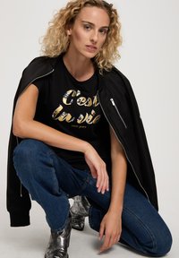 Czarna bomberka noszona na czarnym T-shircie z złotymi i srebrnymi napisami, zestawiona z niebieskimi dżinsami i błyszczącymi srebrnymi botkami za kostkę.