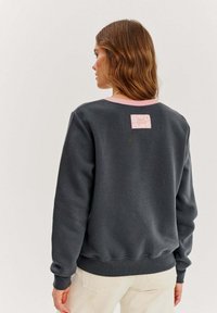 Tmavosivý mikina s ružovým akcentním výstrihom, so malou štítkou s kvetinovým motívom na zadnej strane.