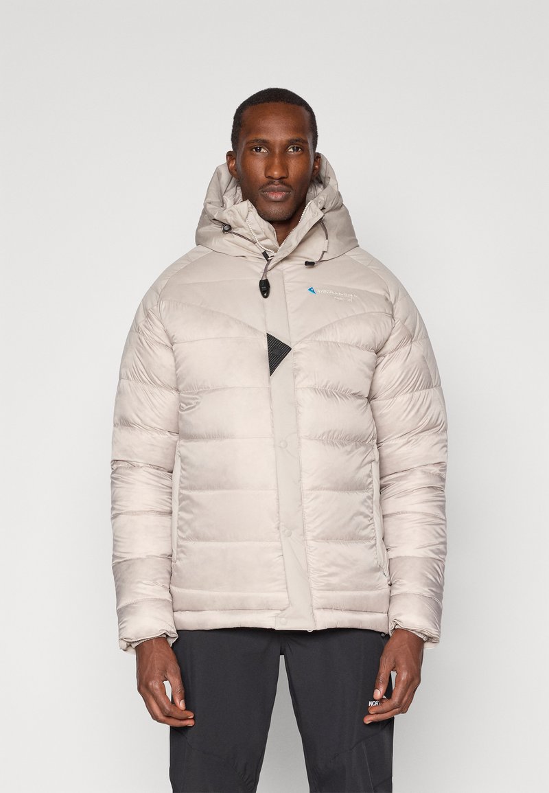 Jachetă puffer de culoare bej deschis, cu glugă. Prezintă un accent triunghiular negru, închidere cu fermoar și quiltare texturată. Material impermeabil.