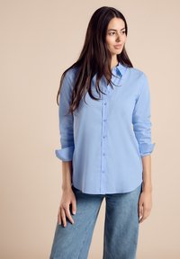 Camisa de botón de color azul claro con cuello, mangas largas y puños enrollados, combinada con jeans de mezclilla de corte holgado. Tejido suave, diseño sencillo.