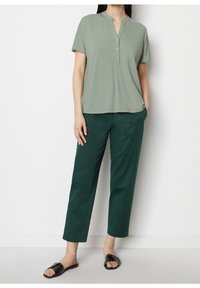 Camiseta de manga corta verde claro con cuello mao y tapeta de botones, combinada con pantalones tipo pinza verde oscuro. Sandalias negras de deslizar completan el conjunto.