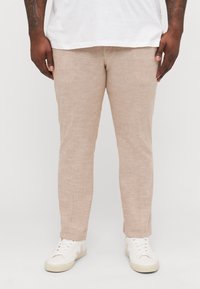 Helle beige, strukturierte Hose mit slim fit. Kombiniert mit weißen Sneakers, die ein dezentes Design und Schnürsenkel haben. Darüber ein schlichtes weißes T-Shirt.