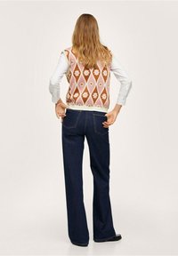 Gestickte Weste mit Blumenmuster in Rosa- und Brauntönen, über einem weißen Shirt getragen, kombiniert mit dunkelblauen Schlagjeans und schwarzen Schuhen.