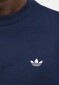 Κοντινό πλάνο ενός ατόμου που φοράει μπλε-σκούφο φούτερ με μικρό λευκό λογότυπο Adidas trefoil στο στήθος.