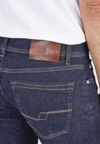 Dunkelblaue Denim-Jeans verfügen über eine glatte Textur, ein braunes Lederetikett mit "HECHTER" und kontrastierende gelbe Nähte an den Taschen.