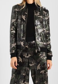 Camo bomberjack met pailletten; zwart en groene kleuren; geribbelde manchetten en zoom; ritssluiting; bijpassende paillettenbroek.