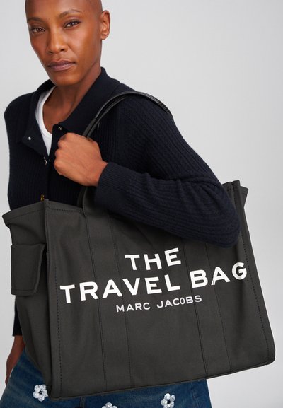 Must mustkunst musta kott valge tekstiga, kus seisab "REISIKOT MARC JACOBS", kahekohaliste käepidemetega ja küljetaskuga.