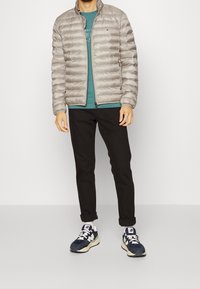 Giacca puffer grigia chiara con cuciture orizzontali, colletto alto e cerniera. Indossata sopra una maglietta teal e pantaloni neri, abbinata a scarpe da ginnastica blu.