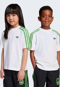 T-shirts brancas com riscas verdes nos ombros, logótipo da Adidas no peito esquerdo. Combinadas com calças pretas com detalhes verdes.