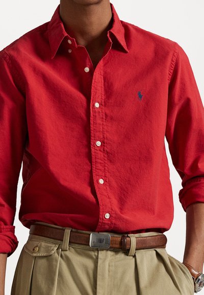 Punasest puhasest puuvillast särk, millel on klassikaline krae, valged nööbid ja väike sinine logo. Kombineeritud khaki pükste ja pruunist vööst.