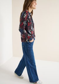 Kolorowy kardigan w wzory z zygzakowatym motywem w kolorach czerwonym, niebieskim i białym, zestawiony z szerokimi niebieskimi dżinsami i białymi trampkami.