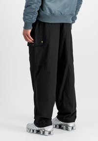 Hombre con pantalones cargo negros con puños ajustables y zapatos deportivos plateados, mostrado desde la cintura hacia abajo sobre un fondo blanco.