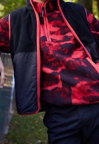 Červený a černý fleecový svetr s výstřihem na zip a černou vrstvou vesty. Má vzor v červených a tmavých odstínech s akcenty na zipu.
