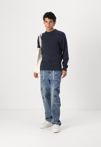 Pulover navy cu manșete côtate, asortat cu blugi cargo de culoare albastru deschis, având buzunare aplicate și margini desfăcute. Adidași albi completează look-ul.