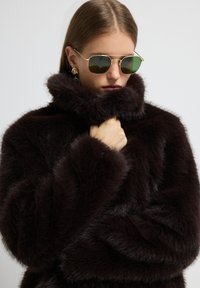 Abrigo de piel sintética marrón oscuro con un cuello alto y una textura gruesa y esponjosa. Los accesorios incluyen gafas de sol con tintado verde y pendientes grandes.