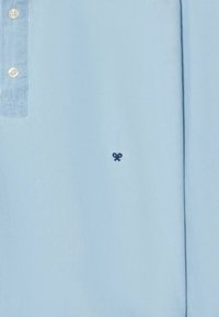 Camisa de manga corta azul claro con cuello abotonado, que presenta un pequeño emblema de tijeras en marino en el lado izquierdo. Textura de tela suave.