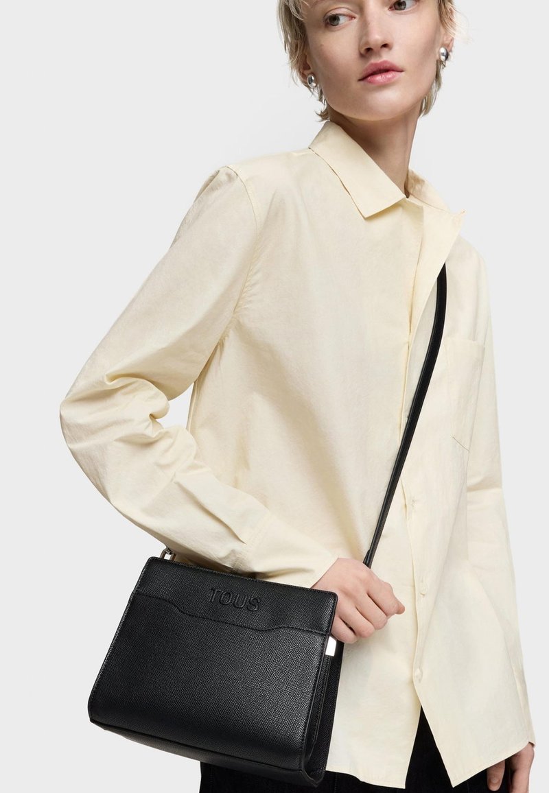 Osoba w jasnej koszuli trzyma czarną torebkę na ramię o teksturowej powierzchni z wytłoczonym logo "TOUS" na jednolitym jasnym tle.