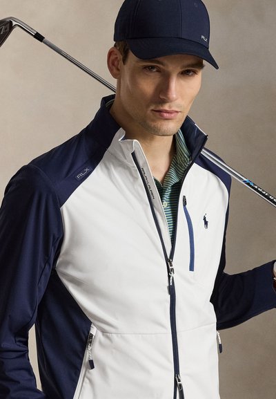 Muž s námořnickou čepicí a bílomodrou golfovou bundou drží golfovou hůl opřenou o rameno, neutrální pozadí.