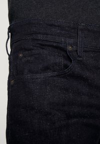 Los jeans de mezclilla oscura presentan un diseño clásico con un corte estrecho, cinco bolsillos y costuras en contraste. La tela tiene una ligera textura.